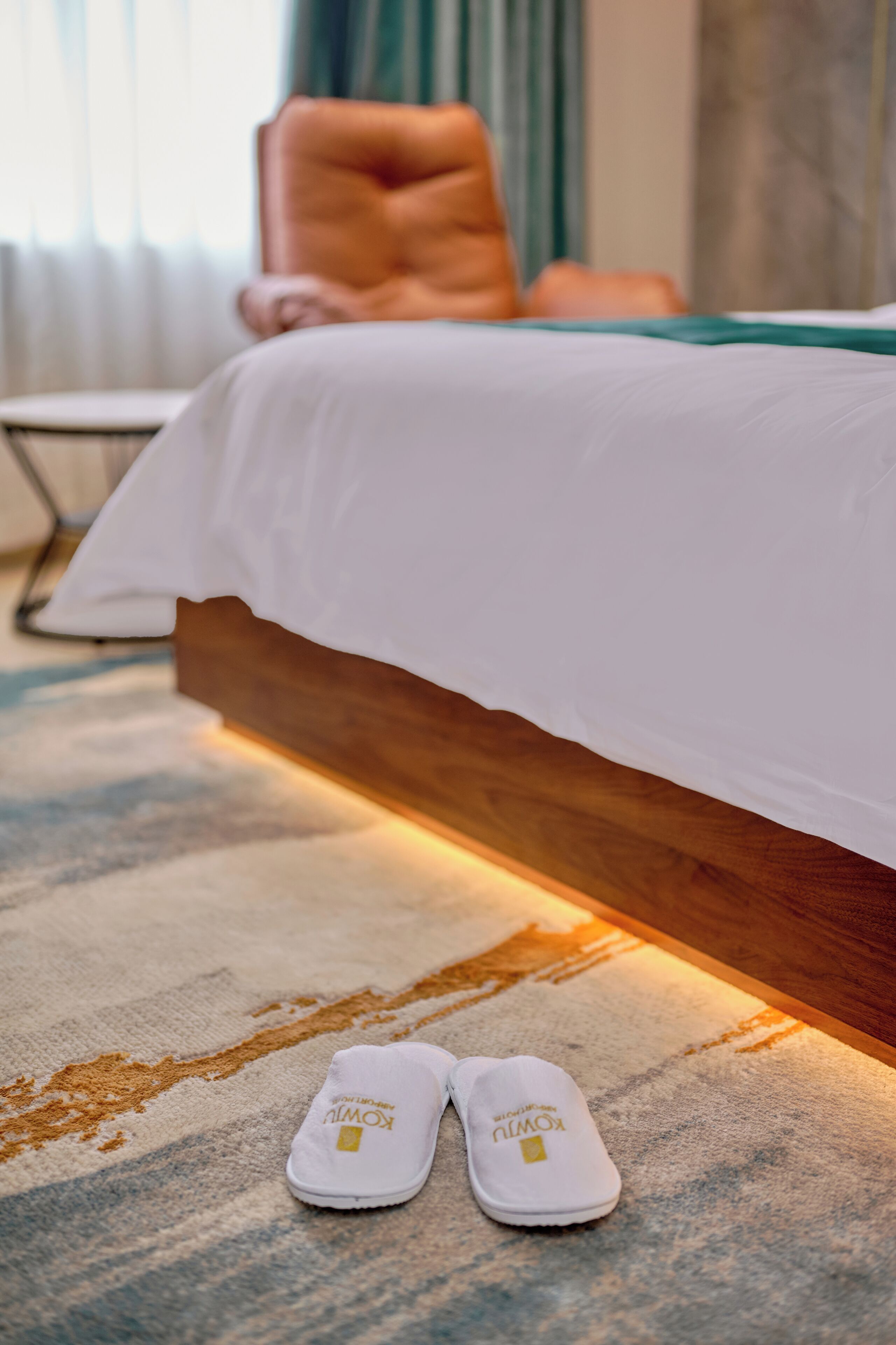 A neatly made bed with a white comforter and a pair of slippers on a patterned carpet.