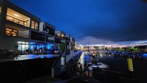 A waterfront marina with boats docked, a modern building with balconies, and outdoor seating areas.