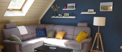 A living room with a grey sectional sofa, a black coffee table, and a lamp on a tripod. There are picture frames on the wall and a window with a view of trees.