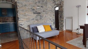 A room with a stone wall, a grey sofa with patterned cushions, a wooden floor, a kitchen area with blue cabinets, and a tall floor lamp. - CasAle sul Lago di Como: serene house with beautiful view of Lake Como  (Blevio)