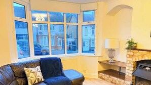 A living room with a leather sofa, a fireplace, and large windows overlooking a street. - Charming Luton Home for Families & Business Stays (Luton)