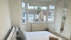 A bedroom with a bed, a radiator, and large windows overlooking a residential area. - Charming Luton Home for Families & Business Stays (Luton)