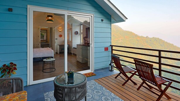 A blue cabin with a balcony, a glass door leading to a bedroom, and a view of a forested hillside. - Mountain Mama Resorts (Narendranagar)