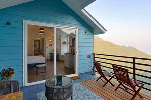 A blue cabin with a balcony, a glass door leading to a bedroom, and a view of a forested hillside. - Mountain Mama Resorts (Narendranagar)