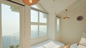 A bedroom with a large bed, two windows, a ceiling fan, and a view of the mountains. - Mountain Mama Resorts (Narendranagar)