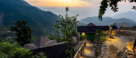 A mountainous landscape with a view of a valley. There are several small buildings with sloped roofs, likely part of a resort or lodge. A wooden fence and a stone wall are visible, with a pathway leading through the area.
