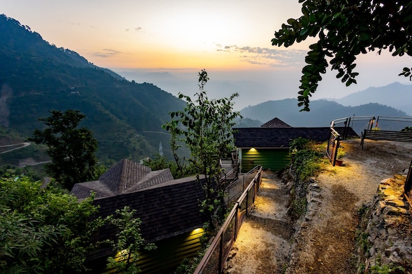 A mountainous landscape with a view of a valley. There are several small buildings with sloped roofs, likely part of a resort or lodge. A wooden fence and a stone wall are visible, with a pathway leading through the area. - Mountain Mama Resorts (Narendranagar)