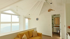 A spacious living room with a large window, a sofa, and a ceiling fan. - Mountain Mama Resorts (Narendranagar)