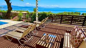 Zona de estar al aire libre con luz solar, sillones reclinables y piscina, con vistas panorámicas al mar y las montañas.