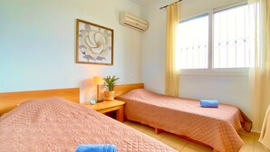 Habitación de hotel con dos camas, una mesita con lámpara, aire acondicionado instalado en la pared y una ventana con cortinas.