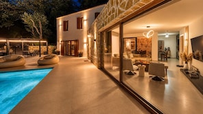 Et moderne hus med swimmingpool, udendørs møbler og et godt oplyst interiør.