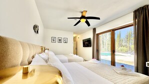 Une chambre d’hôtel avec deux lits, un ventilateur de plafond, une grande fenêtre donnant sur une piscine et des arbres, et des œuvres d’art encadrées accrochées au mur.