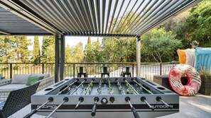 Une table de baby-foot extérieure, dotée d’un cadre en métal, entourée d’un espace de détente abrité et agrémenté de coussins.