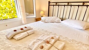 A neatly made bed with white linens and decorative towels with seashells, a bedside lamp, and a window with a view of greenery. - Litsa Beach Maisonette (Acharavi)