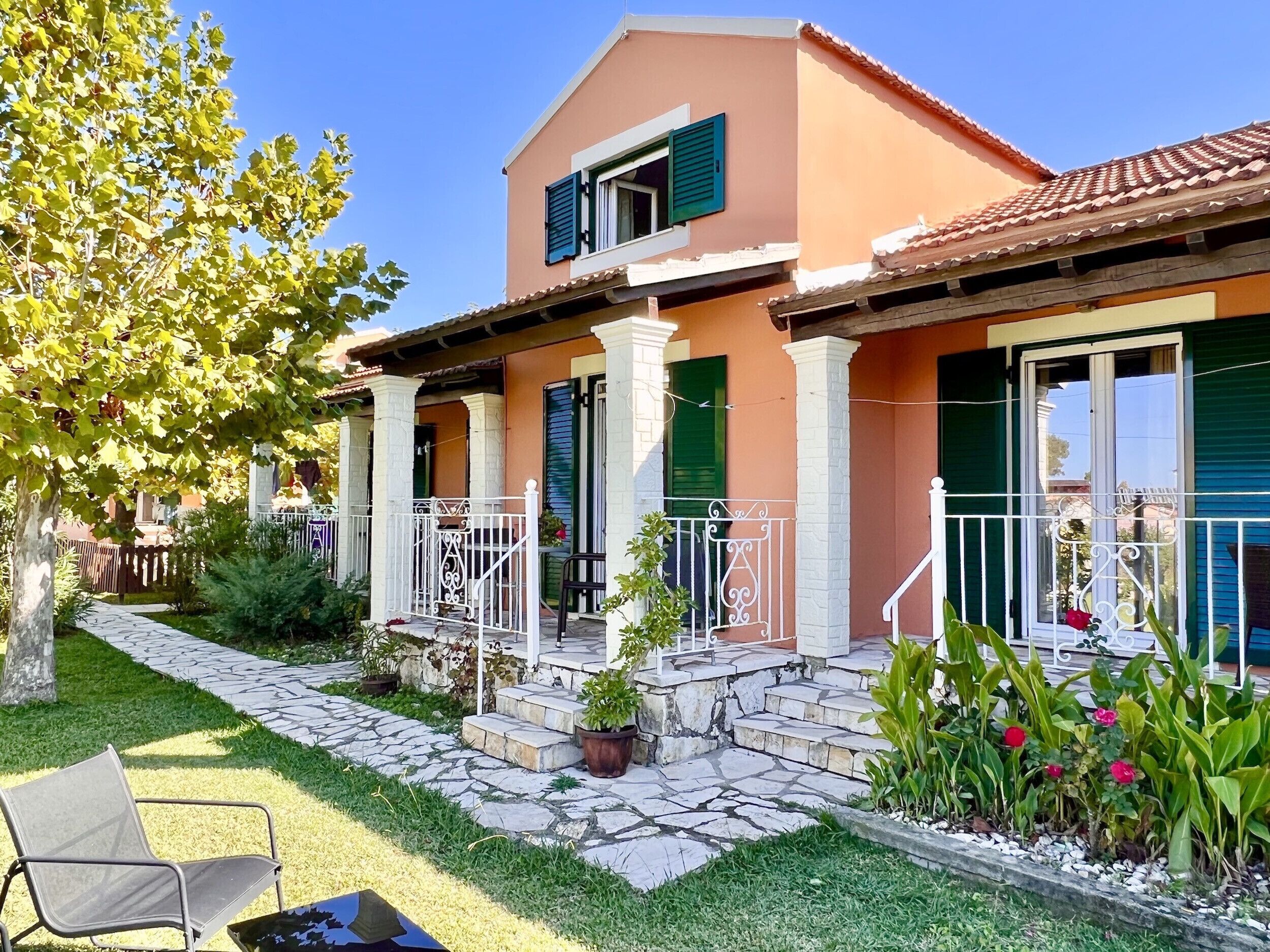 Une maison à deux étages, avec un chemin de pierre menant à l’entrée, un petit porche doté d’une chaise et un jardin agrémenté de plantes et de fleurs en pot.