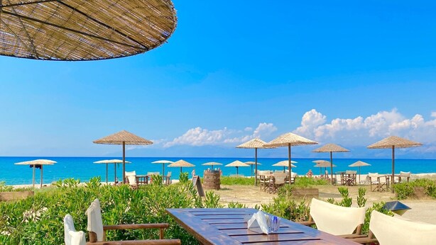 A beachside restaurant with outdoor seating, including wooden tables and chairs, and thatched umbrellas providing shade. - Litsa Beach Maisonette (Acharavi)