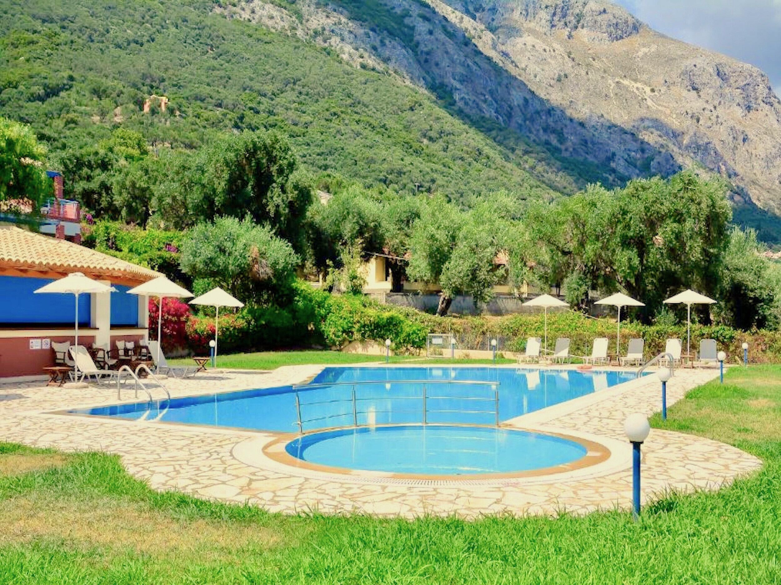 A pool area with lounge chairs and umbrellas, surrounded by lush greenery and mountains.