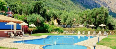 A pool area with lounge chairs and umbrellas, surrounded by lush greenery and mountains.