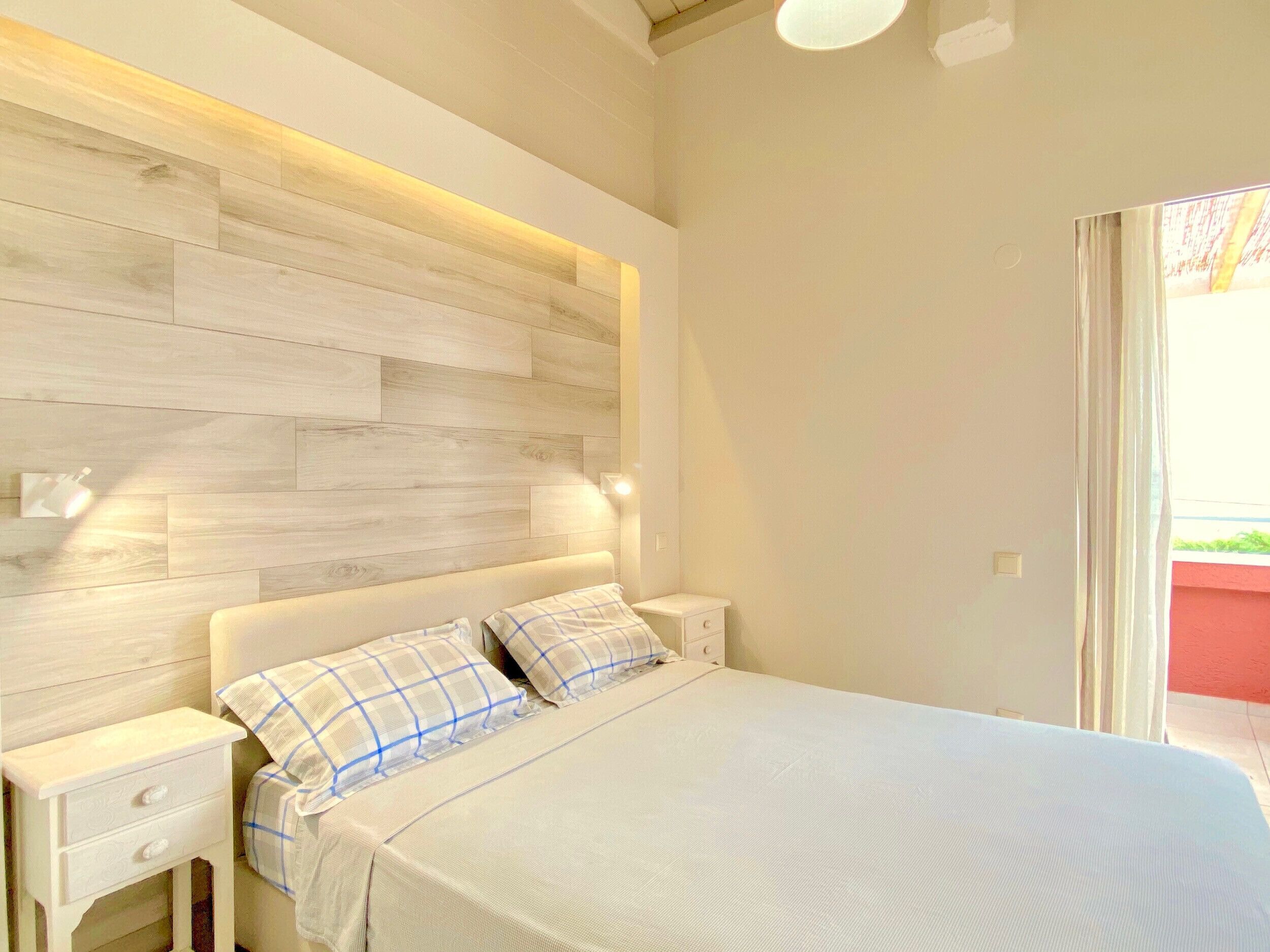 Een slaapkamer met een houten hoofdeinde, een bed met wit beddengoed, een nachtkastje met een lade, en een raam met gordijnen.