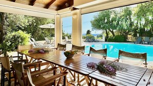 A covered outdoor dining area with a pool view, wooden furniture, and greenery.