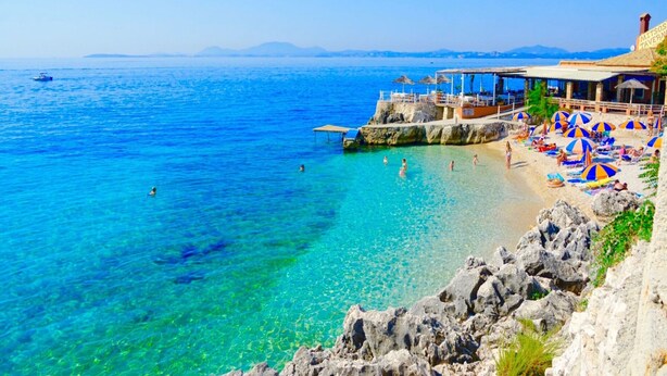 A coastal beach with clear blue waters, rocky shoreline, and a beachside restaurant with umbrellas and seating.