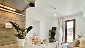 Ein modernes Wohnzimmer mit einem Holzesstisch, einer Couch und einem Sessel. Auf dem Tisch steht eine Pflanze, es gibt eine Deckenleuchte und einen Blick ins Freie durch ein Fenster.