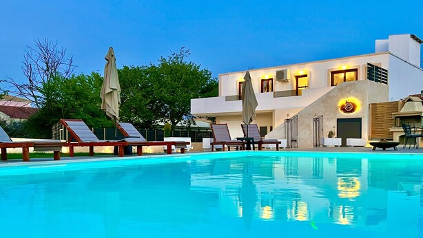 Ein Swimmingpool mit Liegestühlen und Sonnenschirmen, ein modernes Gebäude mit einem Logo und einem wolkenlosen Himmel.
