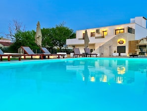 Una piscina con sillas de descanso y sombrillas, un edificio moderno con un logotipo y un cielo despejado.