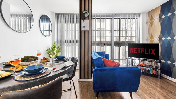 A modern living room with a dining area, a blue sofa, a Netflix TV, and a large window with blinds. - Bullring Lux Suite- Netflix-free Parking-sleeps 6 (Birmingham)