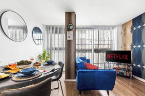 Una sala de estar moderna con comedor, un sofá azul, un televisor Netflix y un ventanal con persianas.