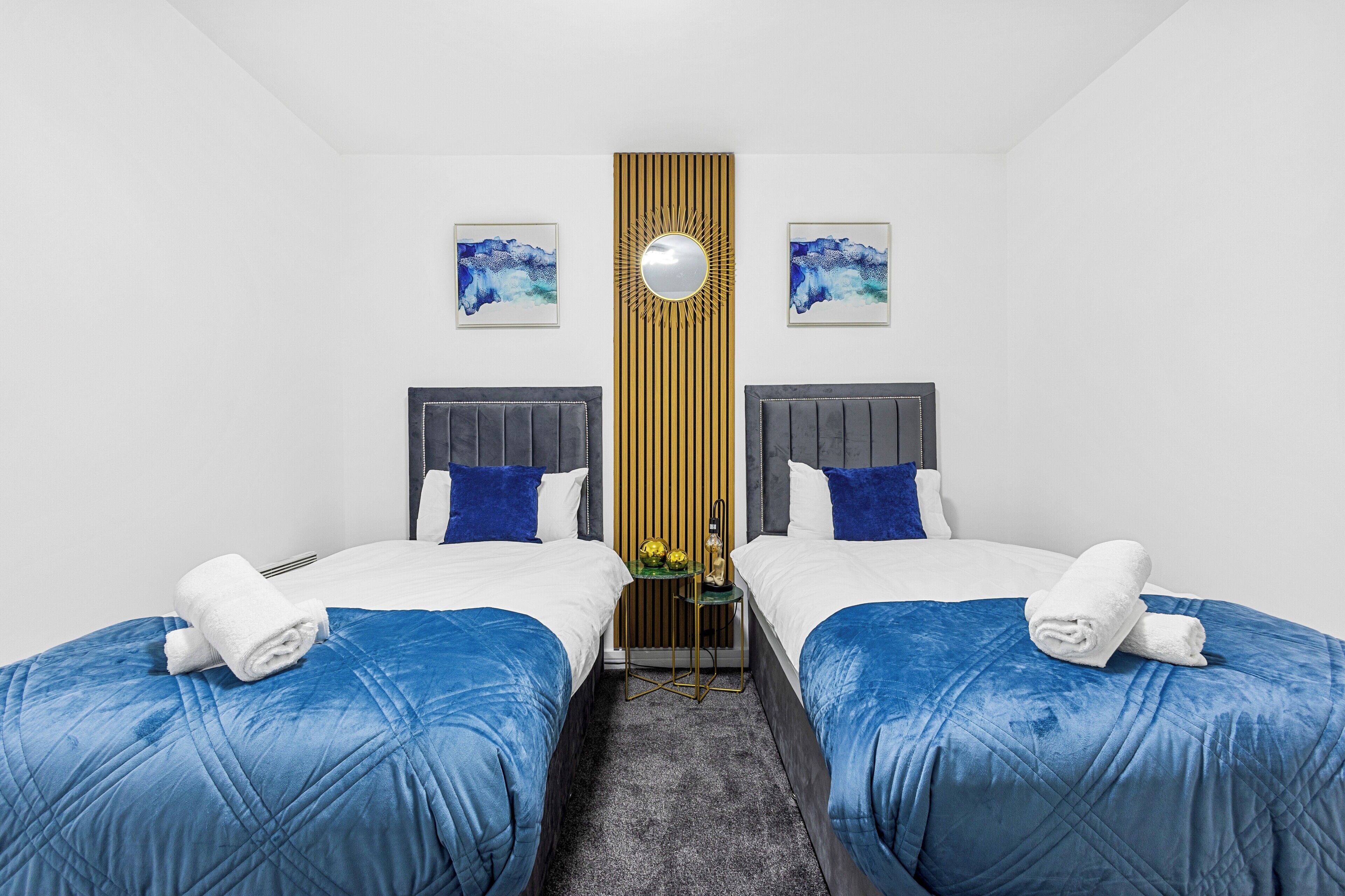 A hotel room with two beds, each with a blue bedspread and white linens, a small side table with a lamp, and a decorative wall panel.