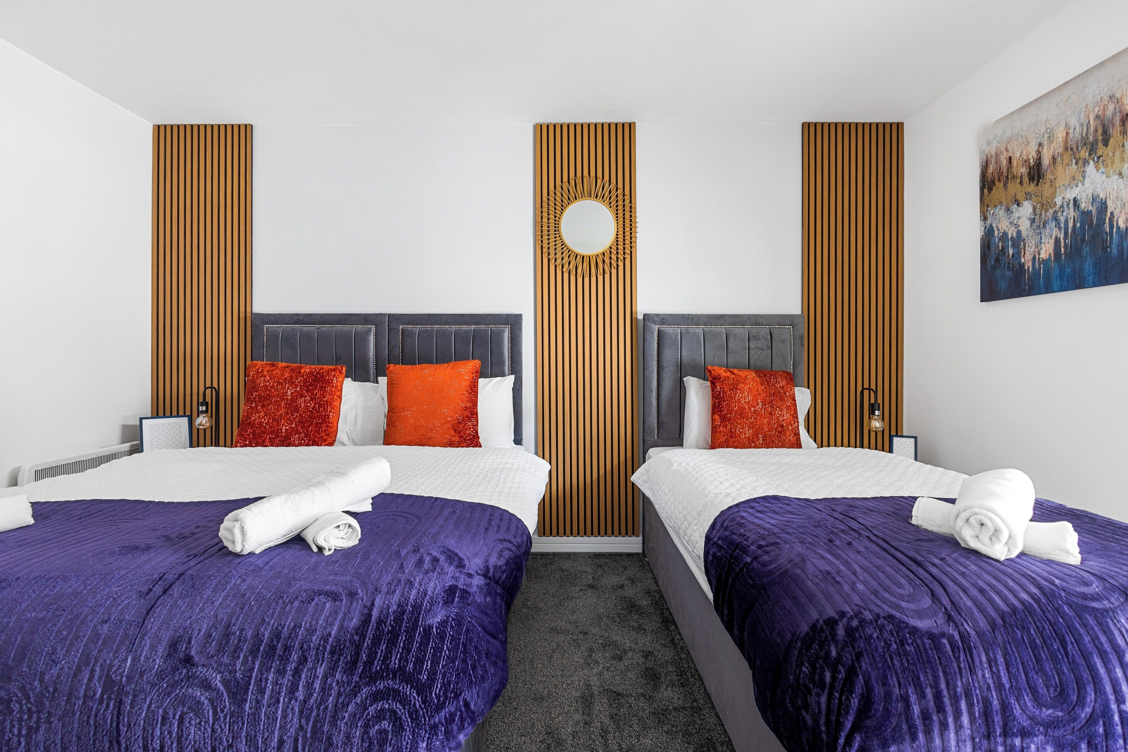 Una camera d'albergo con due letti, ciascuno con copripiumino viola e lenzuola bianche, un pannello di legno a parete, uno specchio rotondo e un quadro appeso al muro.