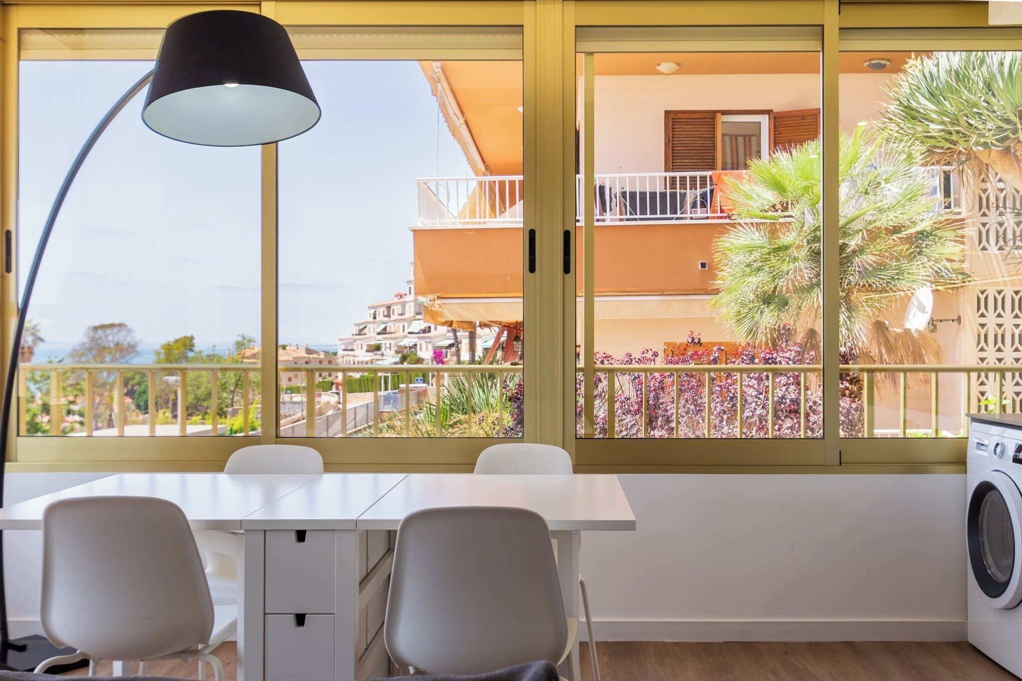 Een kamer met een eettafel, stoelen, een lamp, een wasmachine en uitzicht op een balkon met planten en een gebouw.