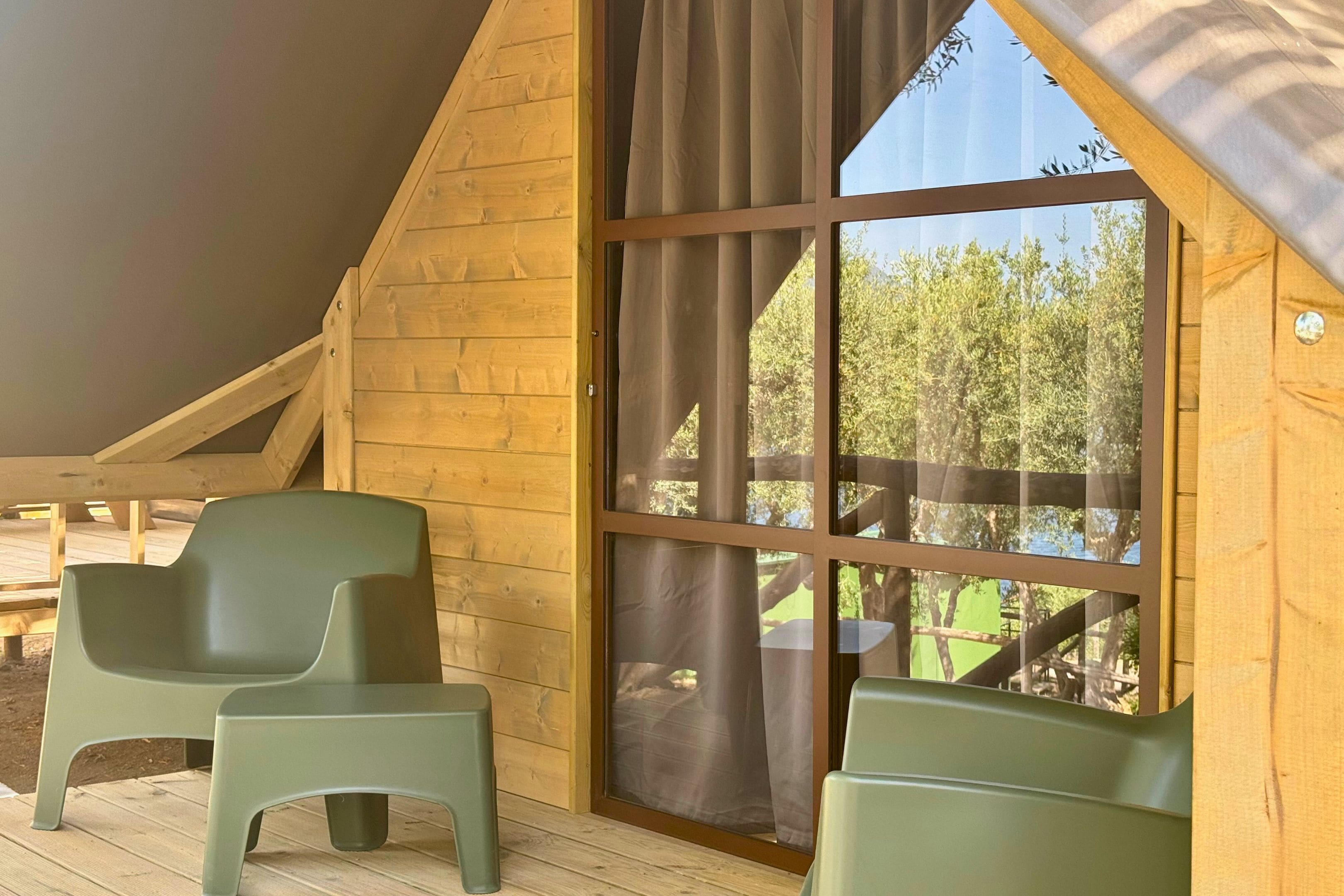 Une cabane en bois avec une grande fenêtre, deux chaises vertes et une terrasse en bois.