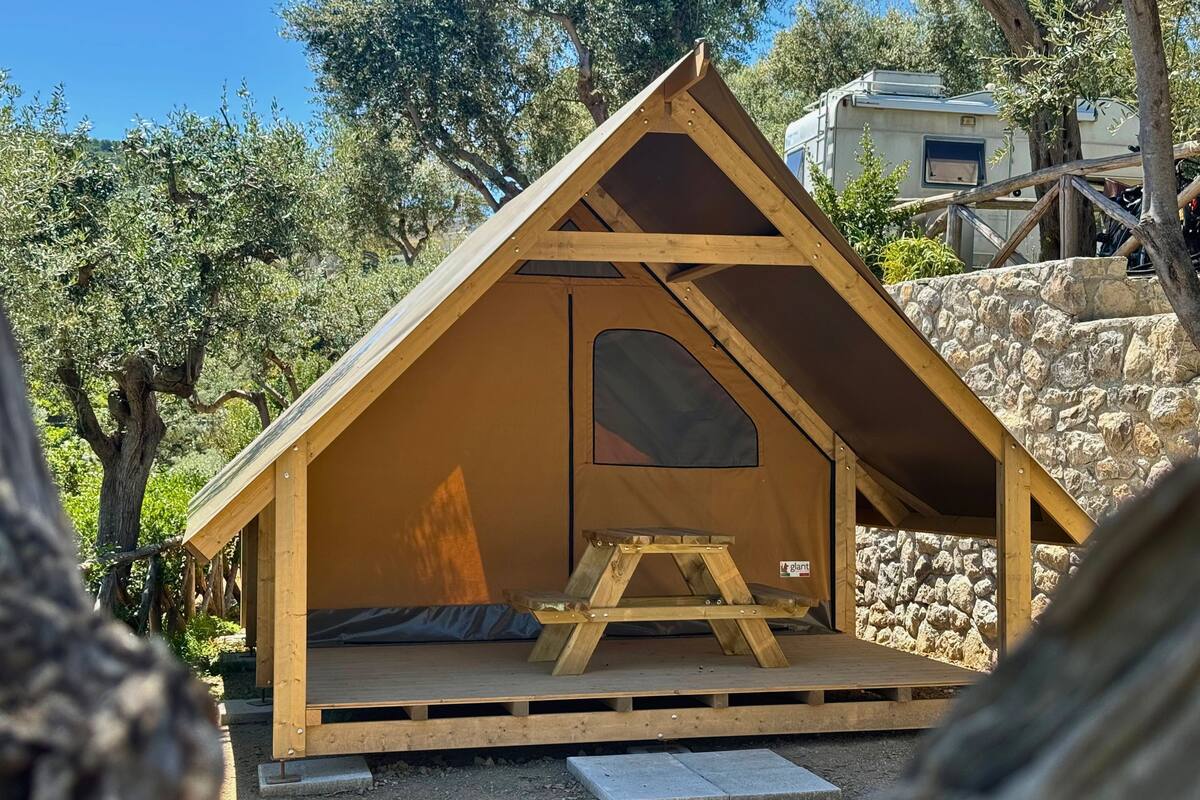 Une petite structure triangulaire en bois, comprenant un espace de couchage et une petite table, entourée d’arbres et d’un mur de pierre.