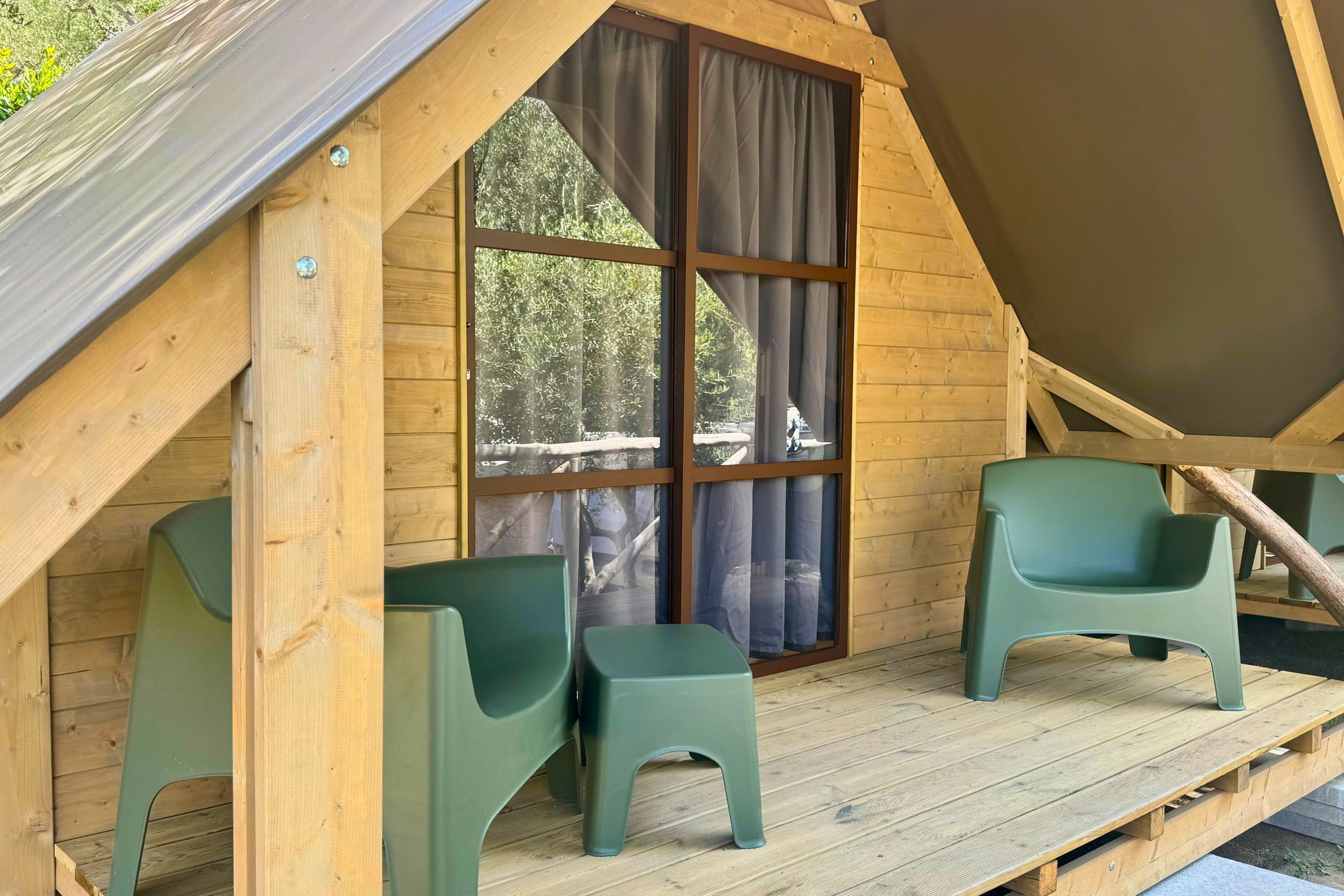 A wooden deck with two green plastic chairs and a large window with curtains.