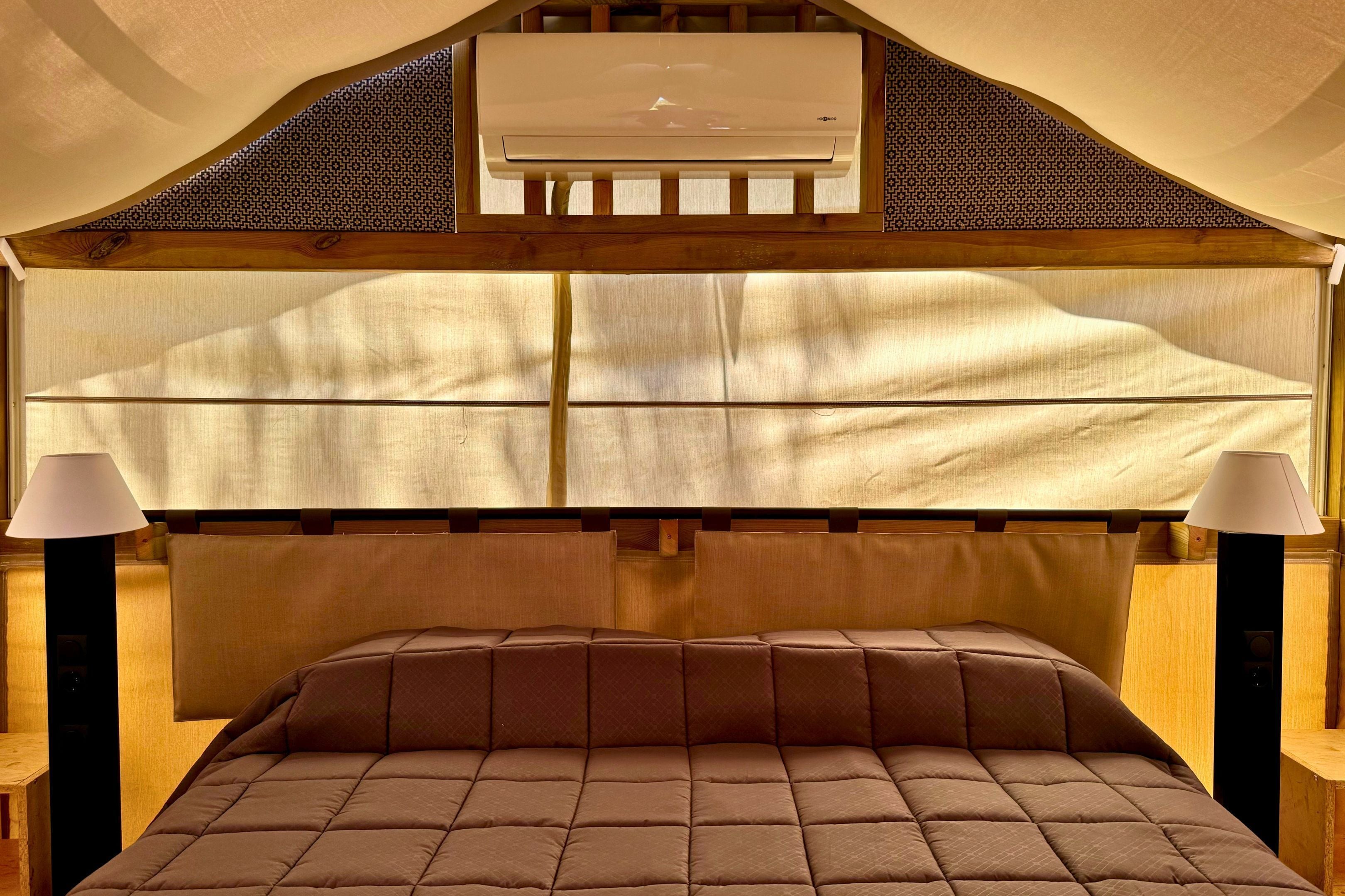 Een slaapkamer met een houten hoofdbord, een bed met een bruin sprei, twee bedlampen en een airconditioning unit boven het hoofdbord.