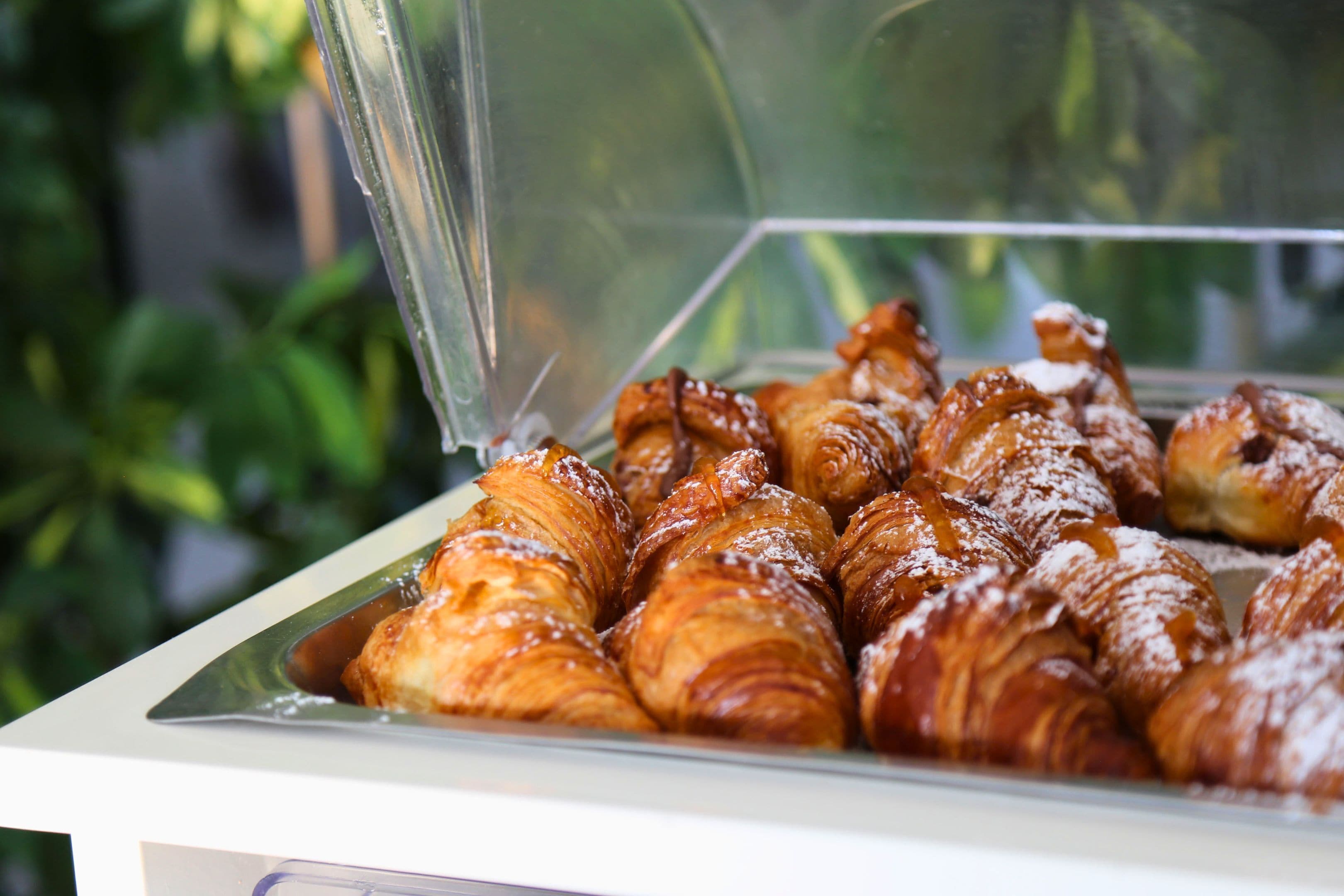 A display case with pastries, including croissants and other baked goods, covered with a clear plastic dome.