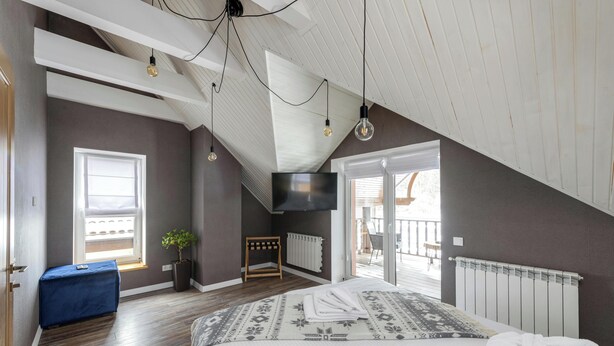 A bedroom with a sloped ceiling, a bed with a patterned bedspread, a TV mounted on the wall, and a window with blinds. - MyTourist (Heumen)