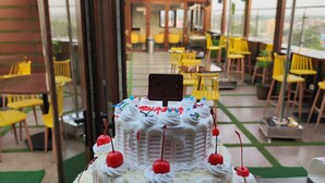 A multi-tiered white cake with cherries on a table in an outdoor dining area with yellow chairs and red umbrellas. - GENX JAMUI (Jamui)