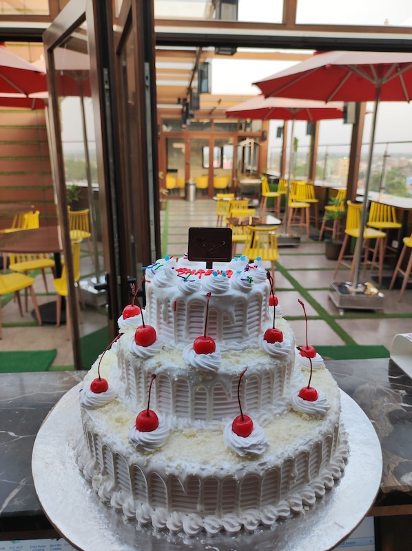A multi-tiered white cake with cherries on a table in an outdoor dining area with yellow chairs and red umbrellas.