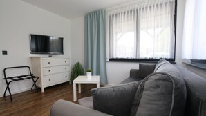 A modern living room with a grey sofa, a white table, a TV on a white cabinet, and a window with sheer curtains. - Ferienhaus Direkt an der Spree (Lübben (Spreewald))