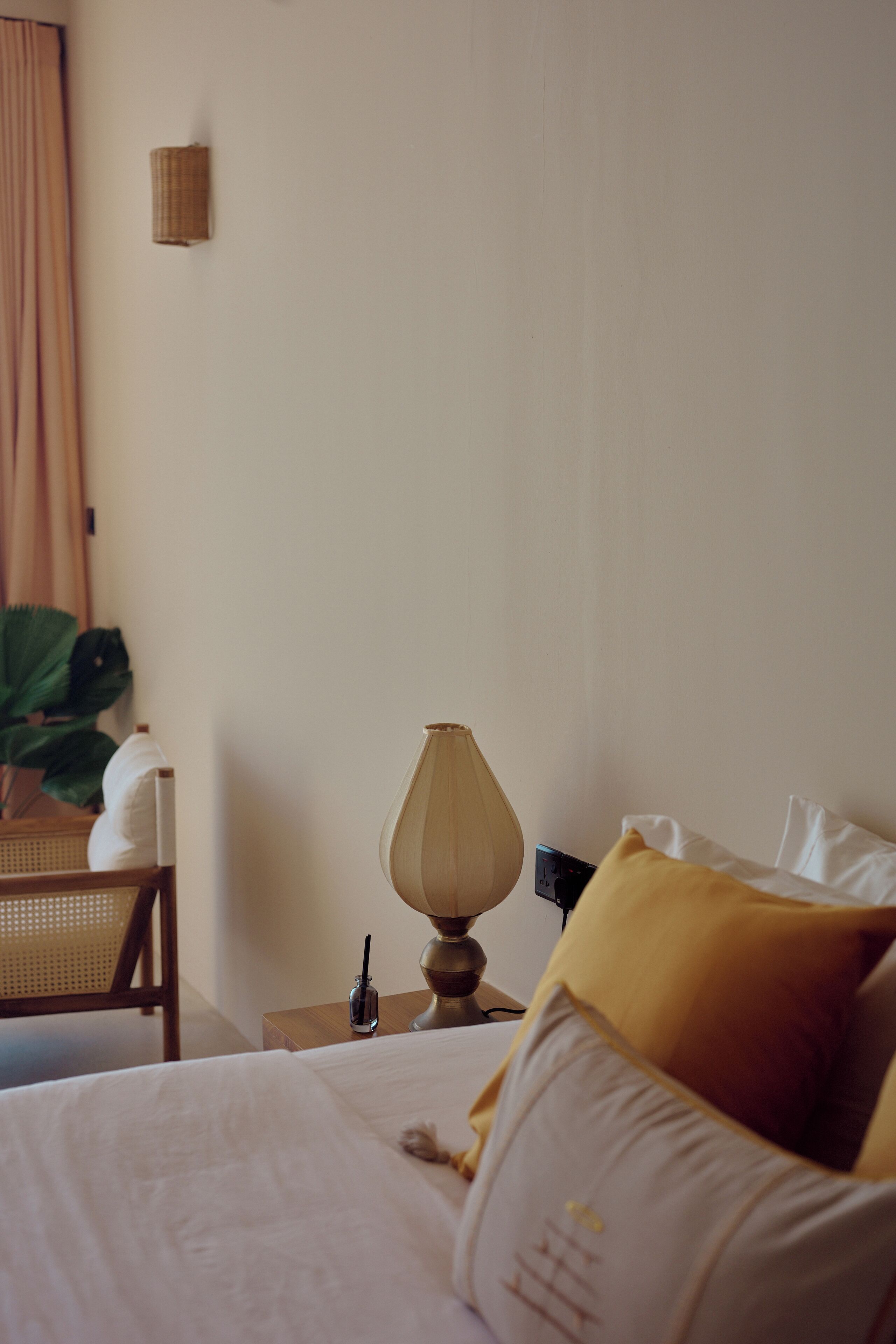 Una cama bien hecha con ropa blanca y una única almohada amarilla, una lámpara de noche, una silla de mimbre y una planta en maceta en una esquina.