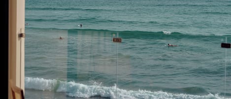 Balcón con sillas y mesa de madera, con vista al mar y surfistas.