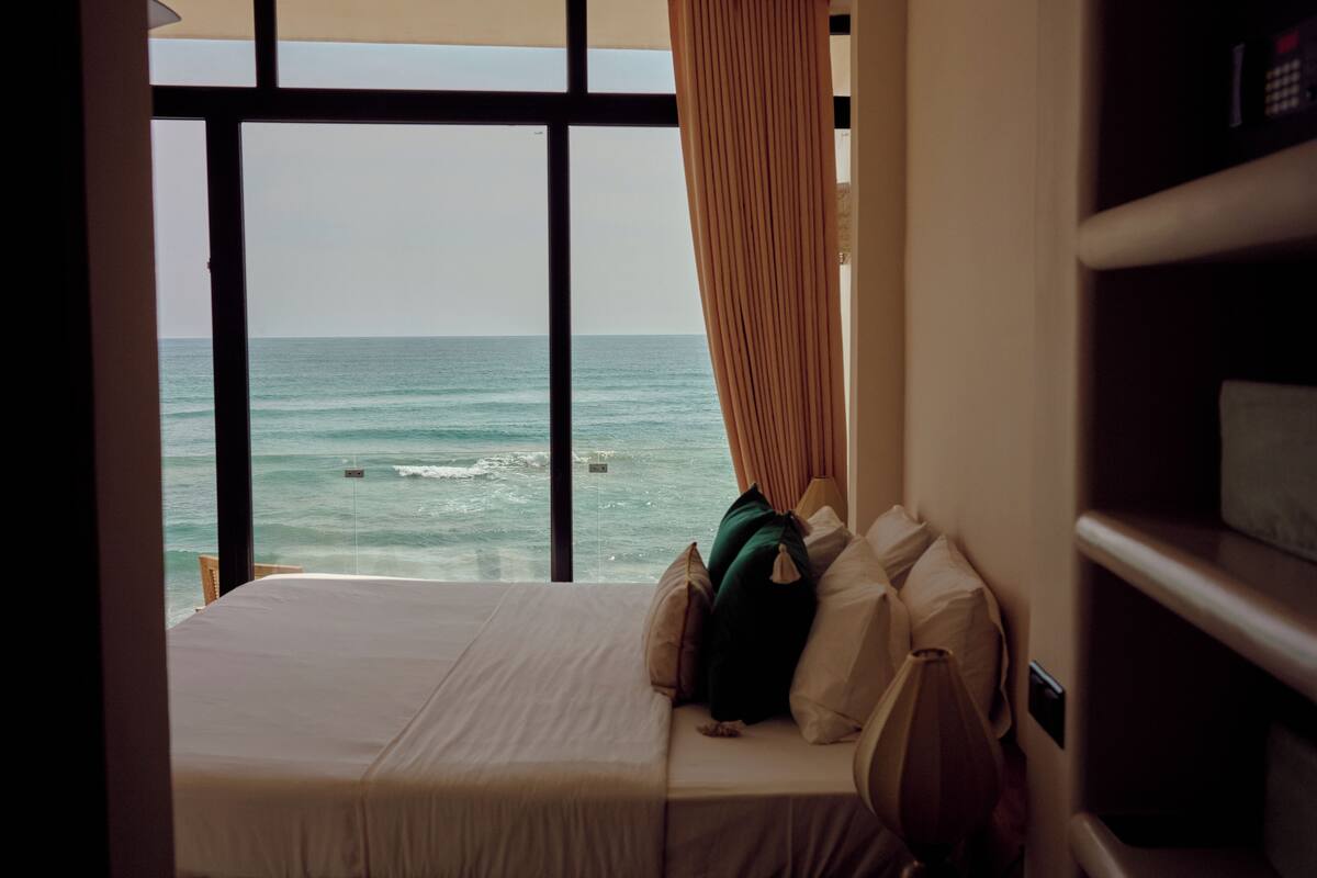 A hotel room with a bed, pillows, and a view of the ocean through large windows.
