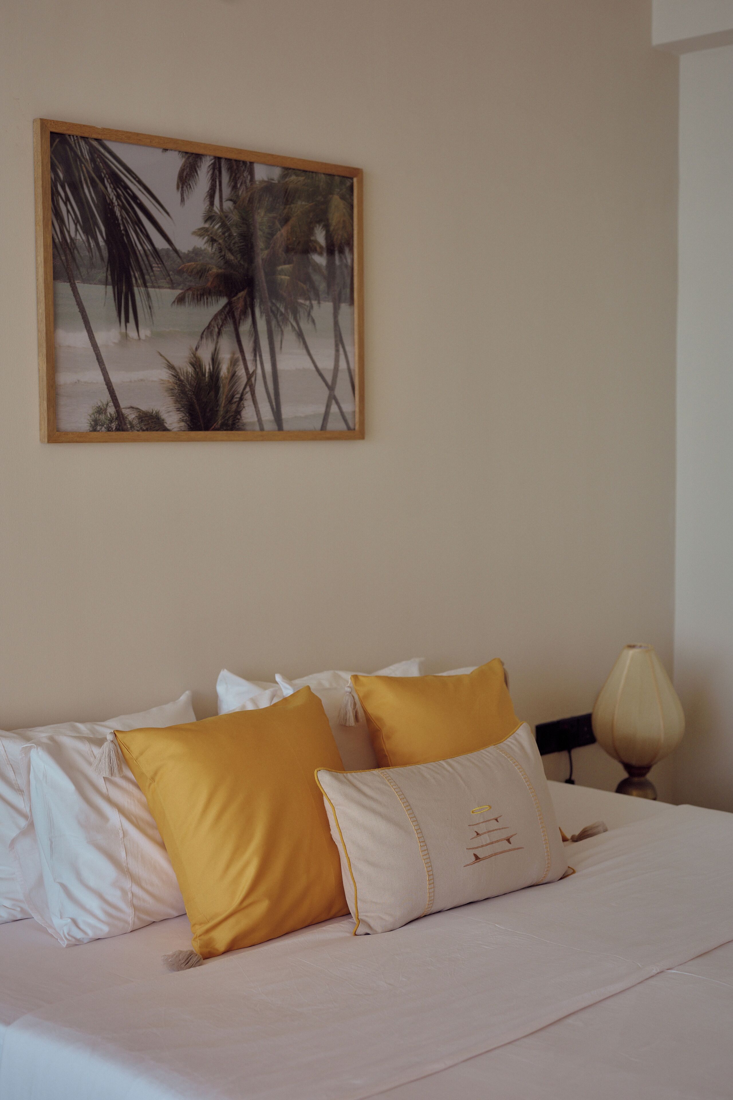 A neatly made bed with white and yellow pillows, a framed picture of palm trees on the wall, and a bedside lamp.
