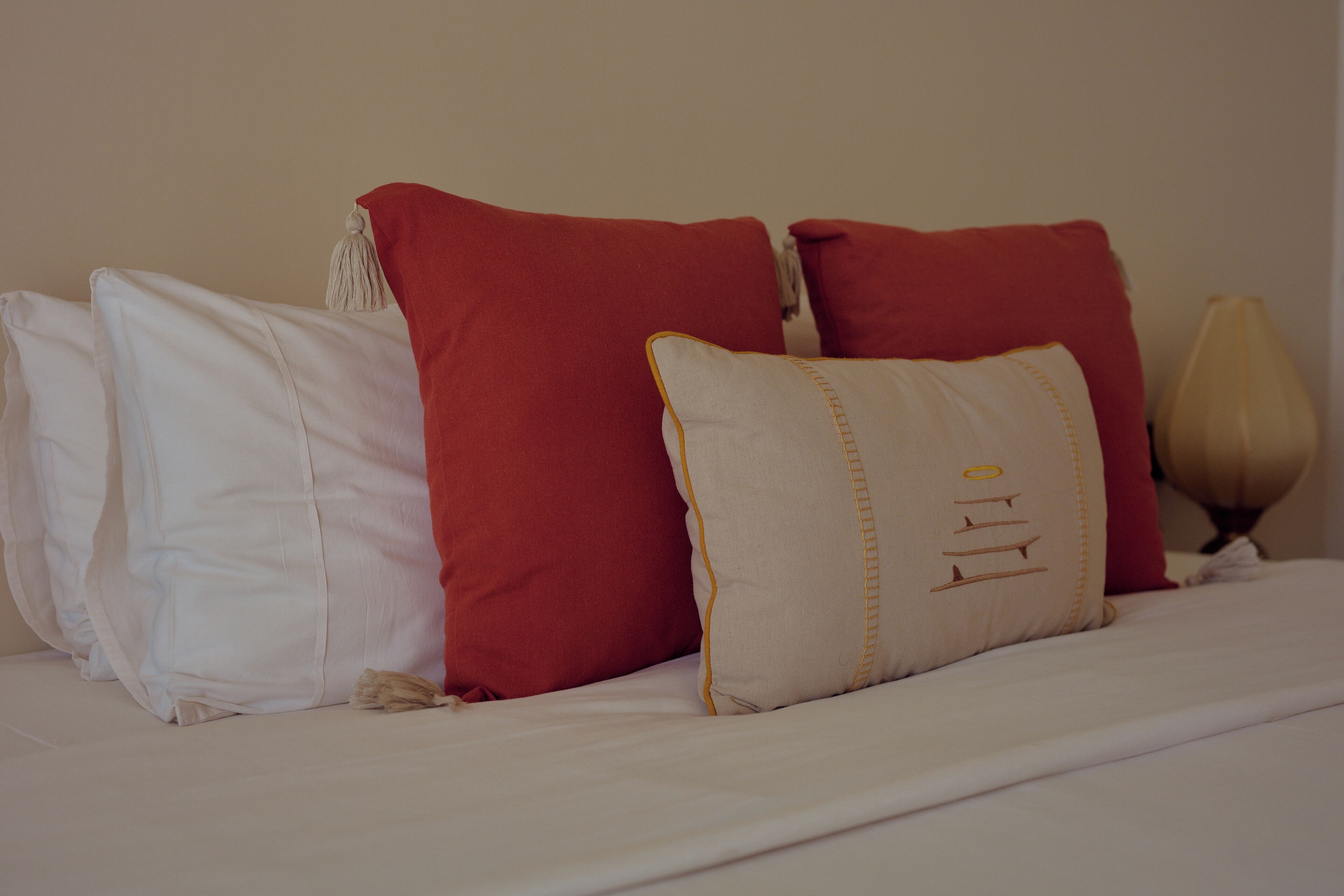 A bed with white and red pillows, a beige pillow with a design, and a lamp on the side.