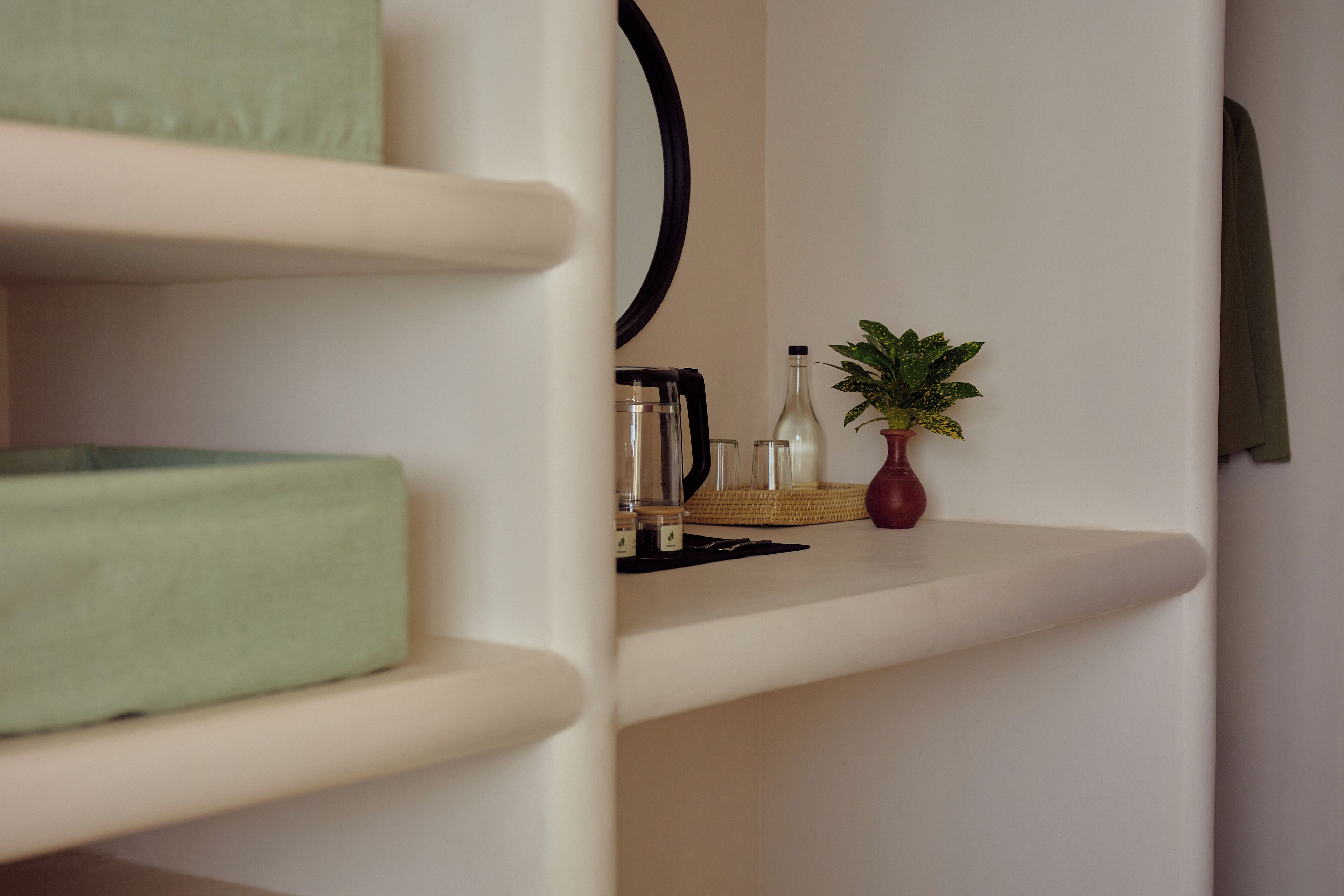Una camera moderna e minimalista con una poltrona imbottita verde, un tavolino con un vaso e una pianta, e uno specchio appeso al muro.