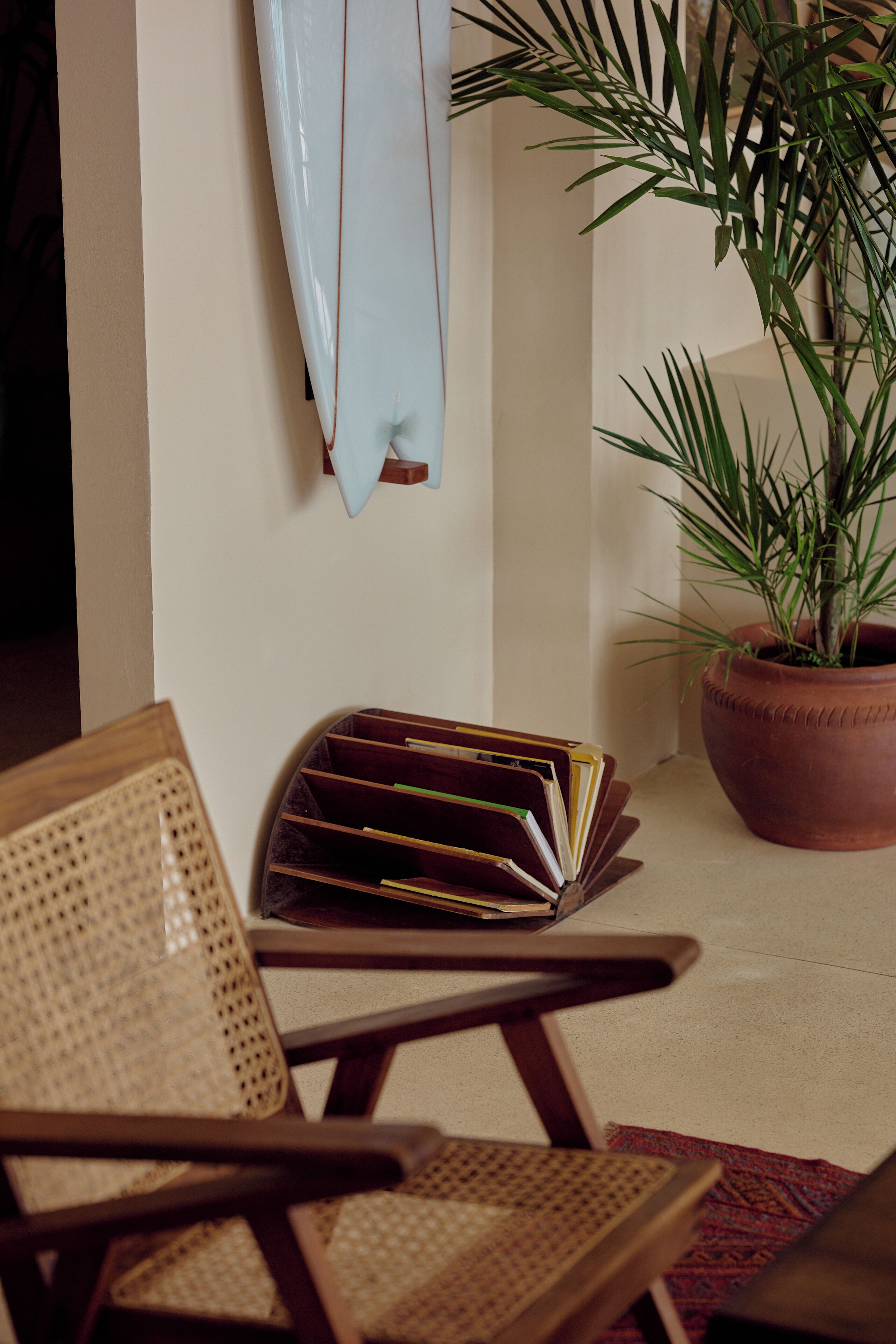 Un coin de pièce avec un fauteuil en osier, une bibliothèque, une plante en pot et une planche de surf appuyée contre le mur.