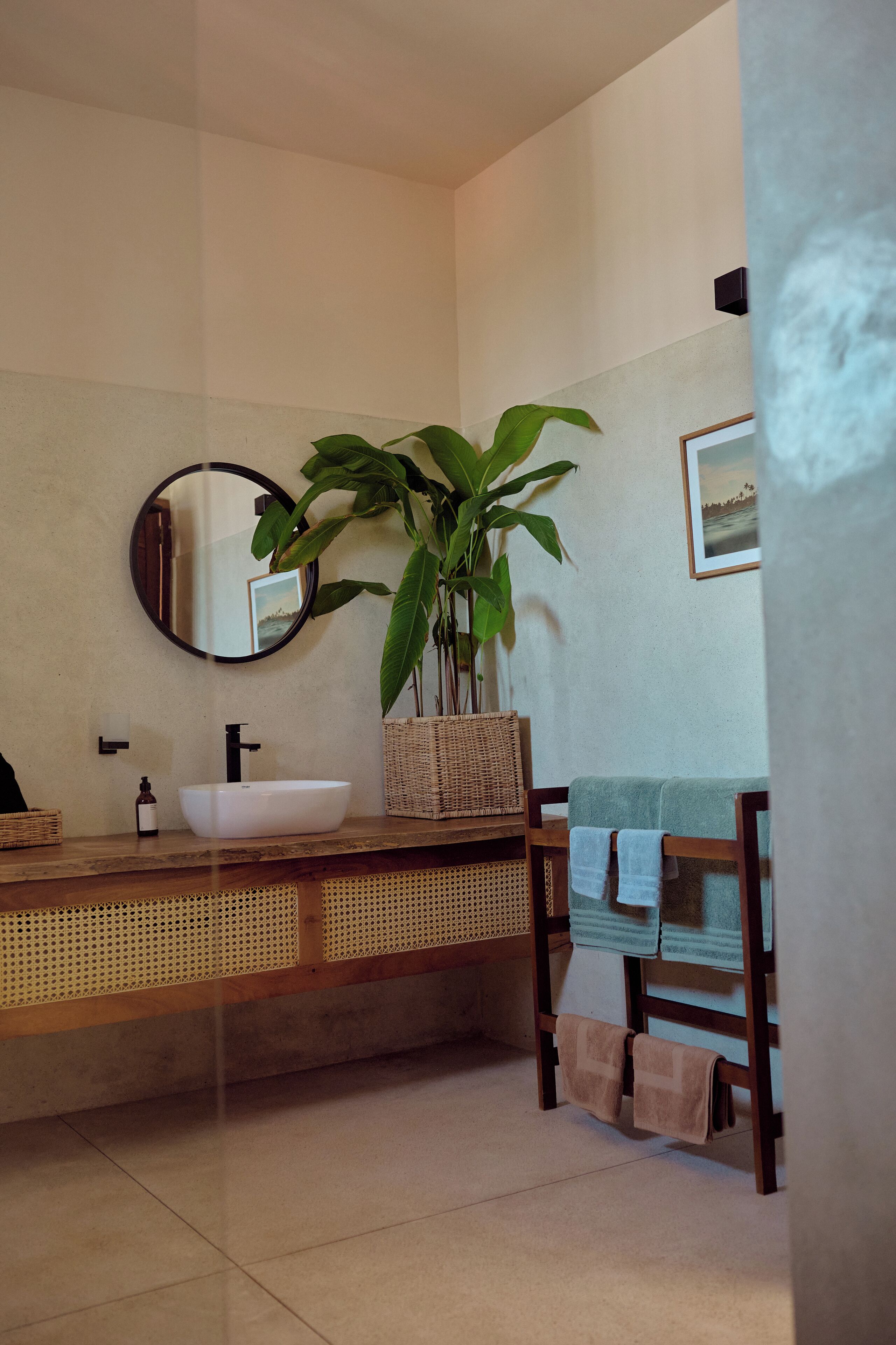 A bathroom with a white sink, a round mirror, a potted plant, and a wooden chair with towels.